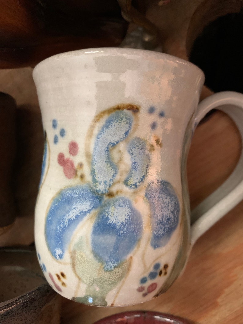 以下が含まれることがあります： 青と緑の花柄が描かれた白い陶器のマグカップ。マグカップは丸みを帯びたボディとハンドルが付いています。
