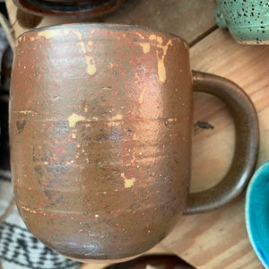 May include: A brown ceramic mug with a speckled glaze and a light brown handle. The mug has a rustic, handmade look.