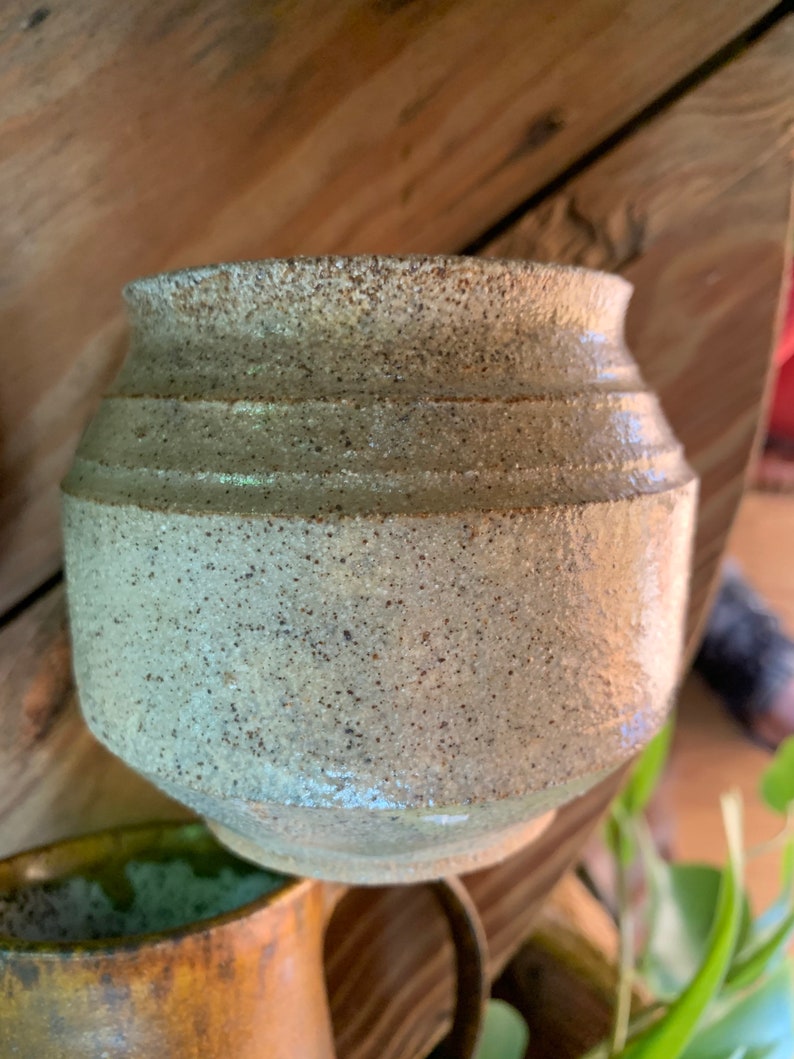 May include: A small, white ceramic pot with a speckled glaze. The pot has a rounded shape and a slightly flared rim.