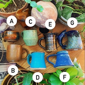 May include: Assortment of eight ceramic mugs in various colours and patterns, including blue, green, brown, and white. The mugs are arranged on a wooden surface with plants in the background.
