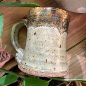 May include: A handmade ceramic mug with a brown and white glaze. The mug has a wide base and a handle.