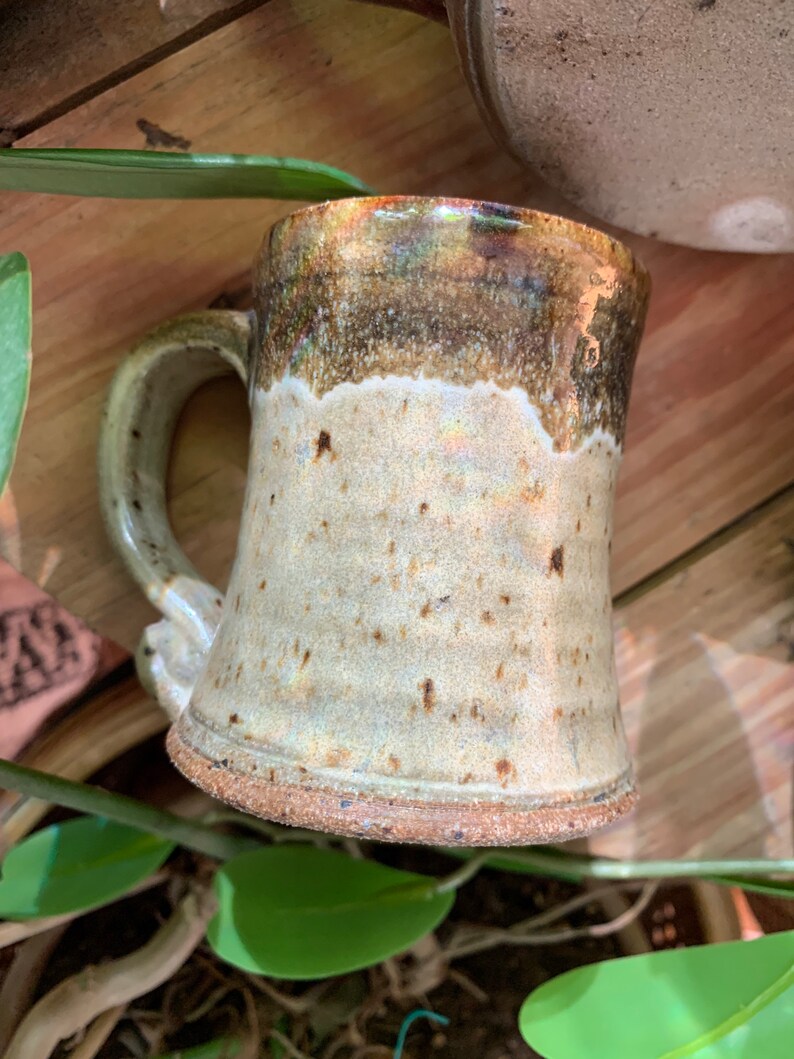 May include: A handmade ceramic mug with a brown and white glaze. The mug has a wide base and a handle.