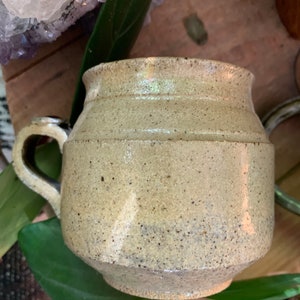 May include: A speckled beige ceramic mug with a rounded body and a handle. The mug has a slightly textured surface.