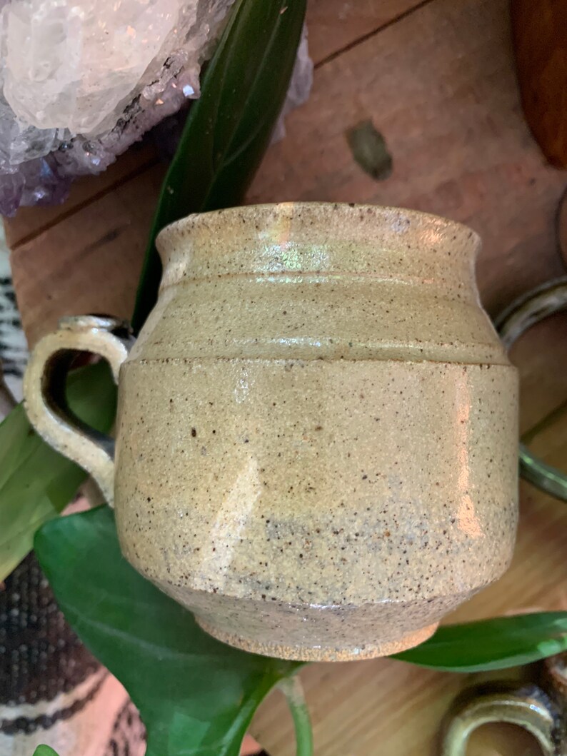 May include: A speckled beige ceramic mug with a rounded body and a handle. The mug has a slightly textured surface.