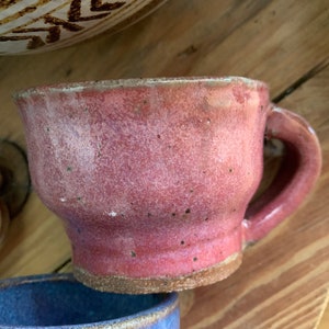 以下が含まれることがあります： 斑点のある釉薬と丸い取っ手がついたピンクの陶器のマグカップ。マグカップは少し傾いていて、素朴で手作り感のある見た目です。