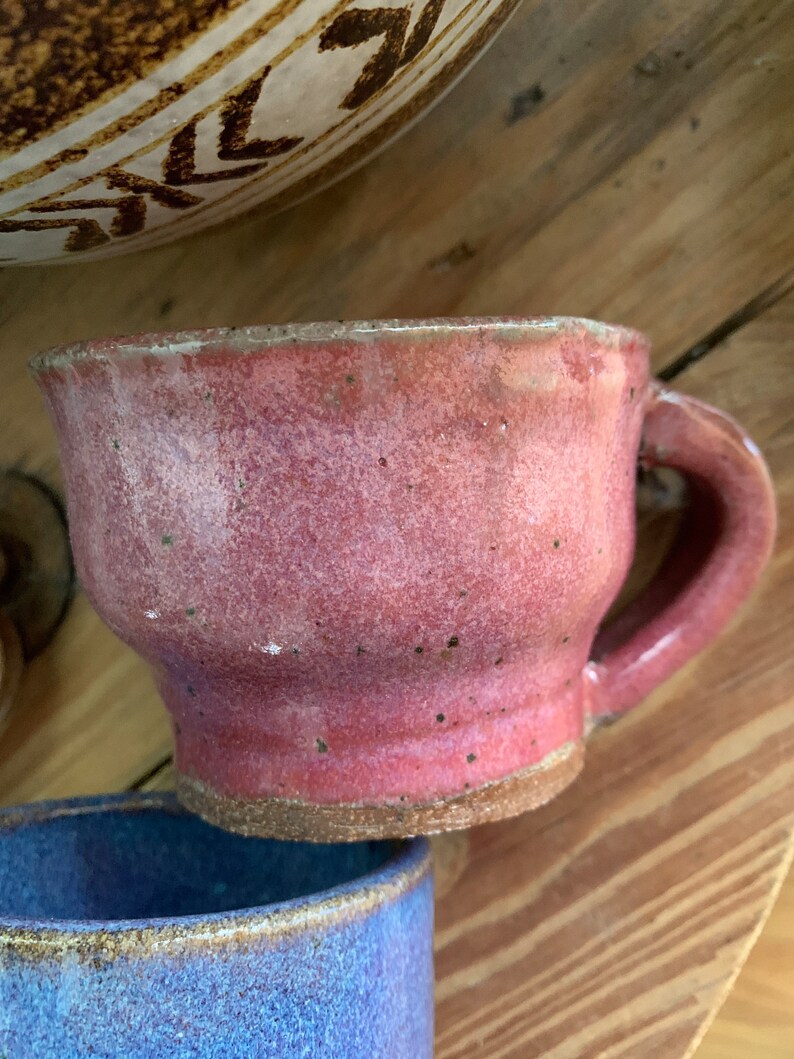 以下が含まれることがあります： 斑点のある釉薬と丸い取っ手がついたピンクの陶器のマグカップ。マグカップは少し傾いていて、素朴で手作り感のある見た目です。