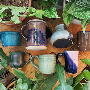 May include: A collection of seven handmade ceramic mugs and a vase in various colours and designs. The mugs are all different shapes and sizes, and some have handles. The vase is a light blue colour with a white stripe around the middle. The mugs and vase are arranged on a wooden surface.