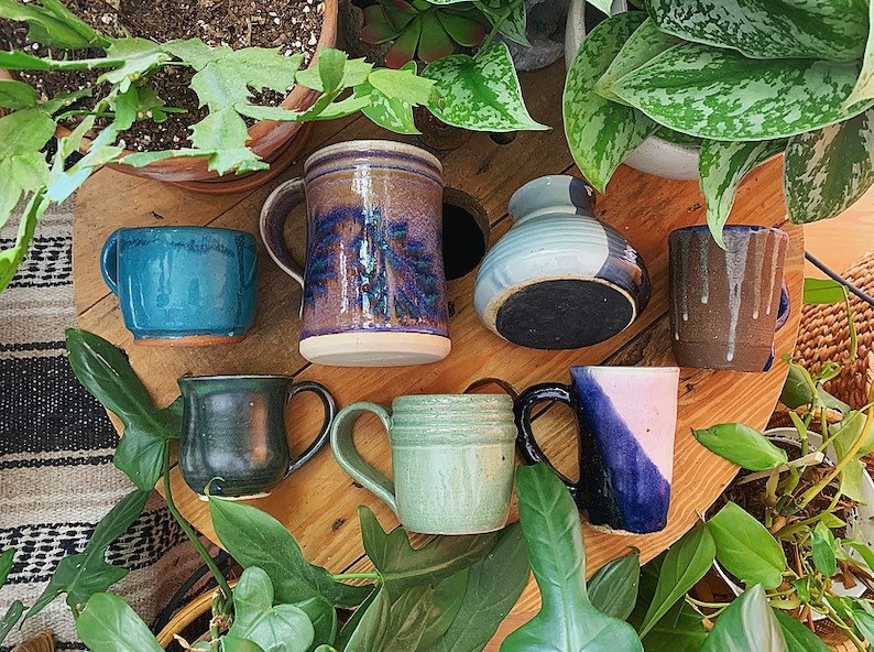 May include: A collection of seven handmade ceramic mugs and a vase in various colours and designs. The mugs are all different shapes and sizes, and some have handles. The vase is a light blue colour with a white stripe around the middle. The mugs and vase are arranged on a wooden surface.