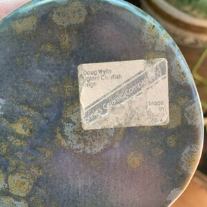 May include: A close-up of the underside of a ceramic glazed blue and green fish sculpture. The base of the sculpture has a white sticker with the text "Doug Wylie Original Clayfish Design Connie's Ceramic Company, NY Made in NY".