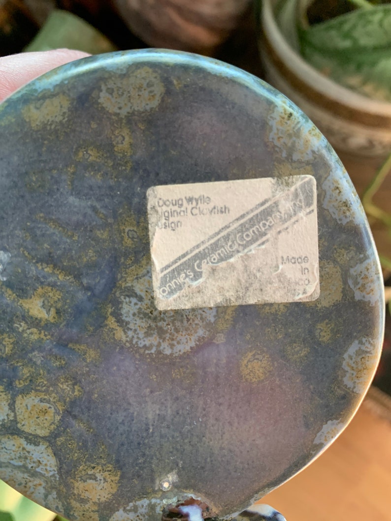 May include: A close-up of the underside of a ceramic glazed blue and green fish sculpture. The base of the sculpture has a white sticker with the text "Doug Wylie Original Clayfish Design Connie's Ceramic Company, NY Made in NY".