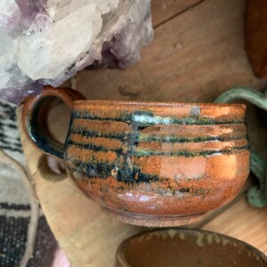 May include: A brown ceramic mug with a black and green glaze. The mug has a wide, rounded body and a handle.