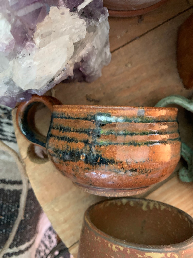 May include: A brown ceramic mug with a black and green glaze. The mug has a wide, rounded body and a handle.