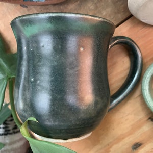 May include: A dark green ceramic mug with a rounded body and a handle. The mug has a speckled glaze and a slightly uneven surface.
