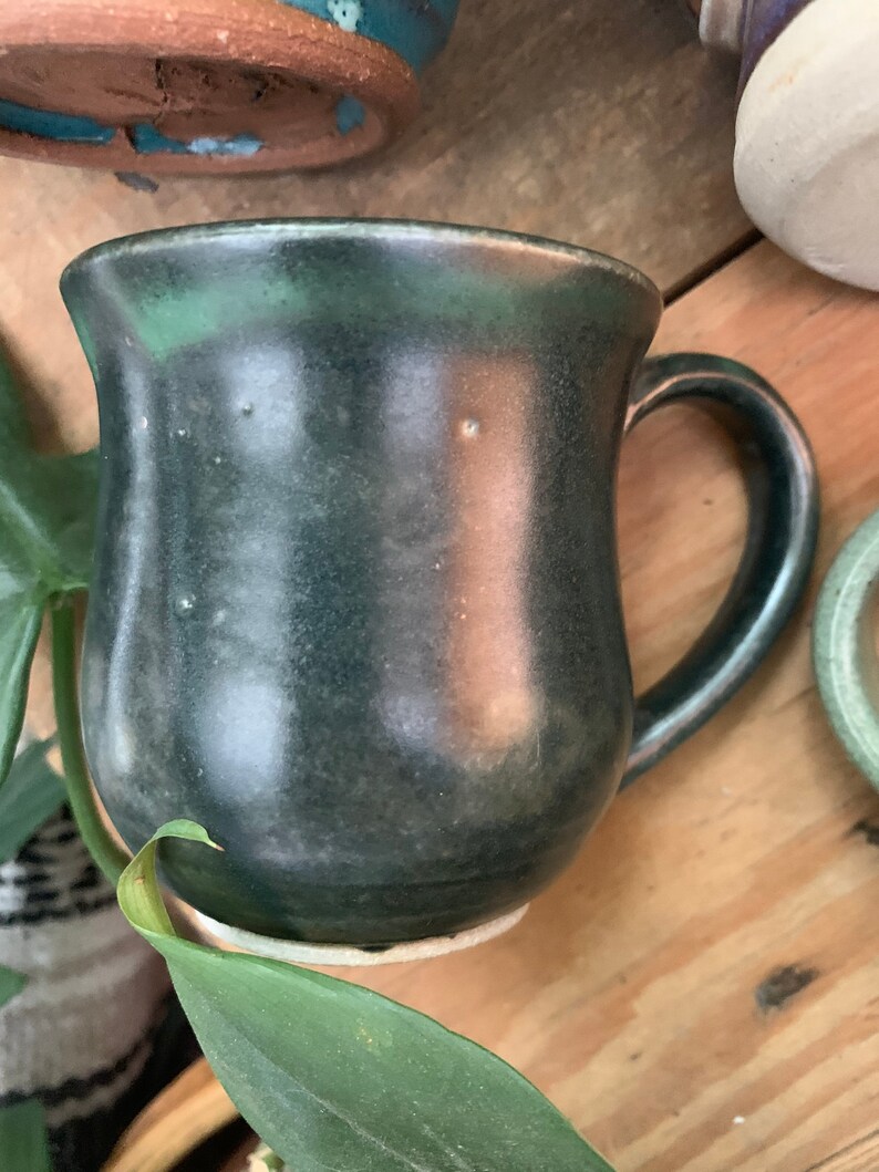 May include: A dark green ceramic mug with a rounded body and a handle. The mug has a speckled glaze and a slightly uneven surface.