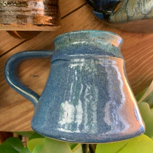 May include: A blue ceramic mug with a speckled glaze and a rounded handle. The mug has a light blue glaze with a darker blue glaze around the rim.