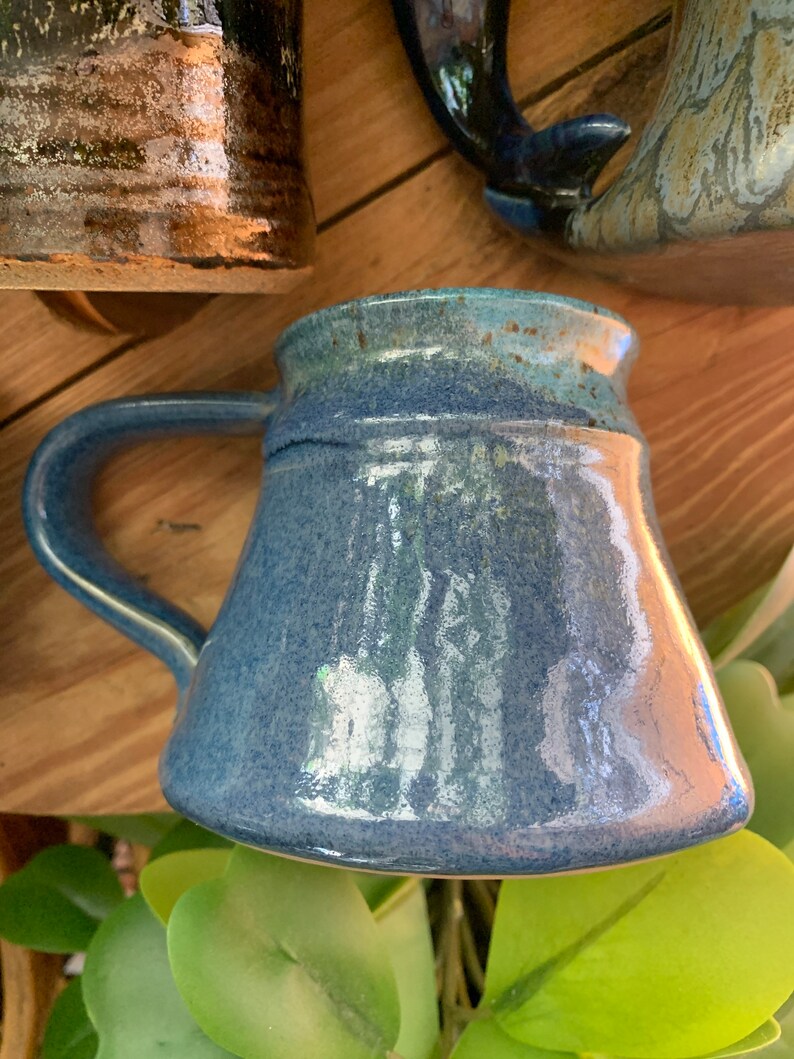 May include: A blue ceramic mug with a speckled glaze and a rounded handle. The mug has a light blue glaze with a darker blue glaze around the rim.