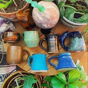 May include: A collection of seven handmade ceramic mugs in various colours and patterns, including blue, green, brown, and white. The mugs are arranged on a wooden table with plants and a decorative stone sphere.