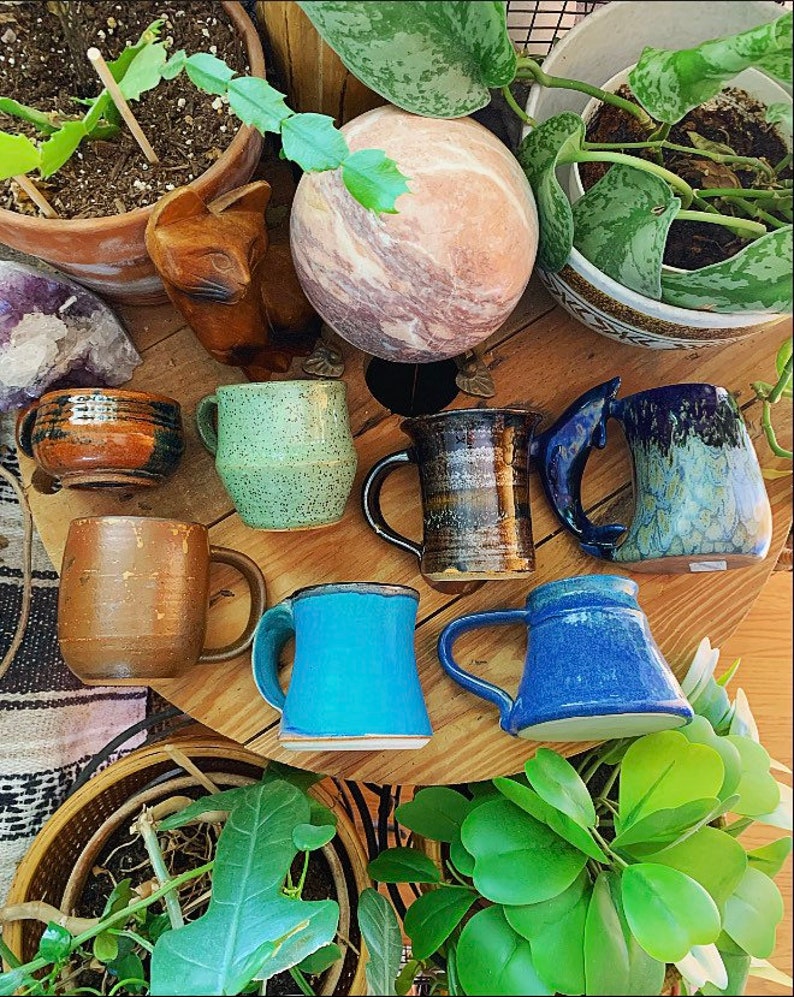 May include: A collection of seven handmade ceramic mugs in various colours and patterns, including blue, green, brown, and white. The mugs are arranged on a wooden table with plants and a decorative stone sphere.