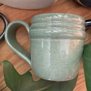 May include: A light green ceramic mug with a rounded handle and three raised bands around the top. The mug has a speckled glaze.
