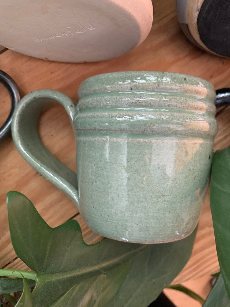 May include: A light green ceramic mug with a rounded handle and three raised bands around the top. The mug has a speckled glaze.