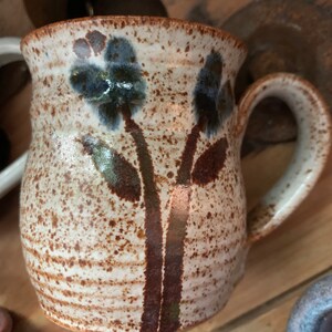 以下が含まれることがあります： 茶色の斑点のある釉薬と青い花がついた茶色の花の模様が施された白い陶器のマグカップ。マグカップは丸みを帯びたボディとハンドルが付いています。
