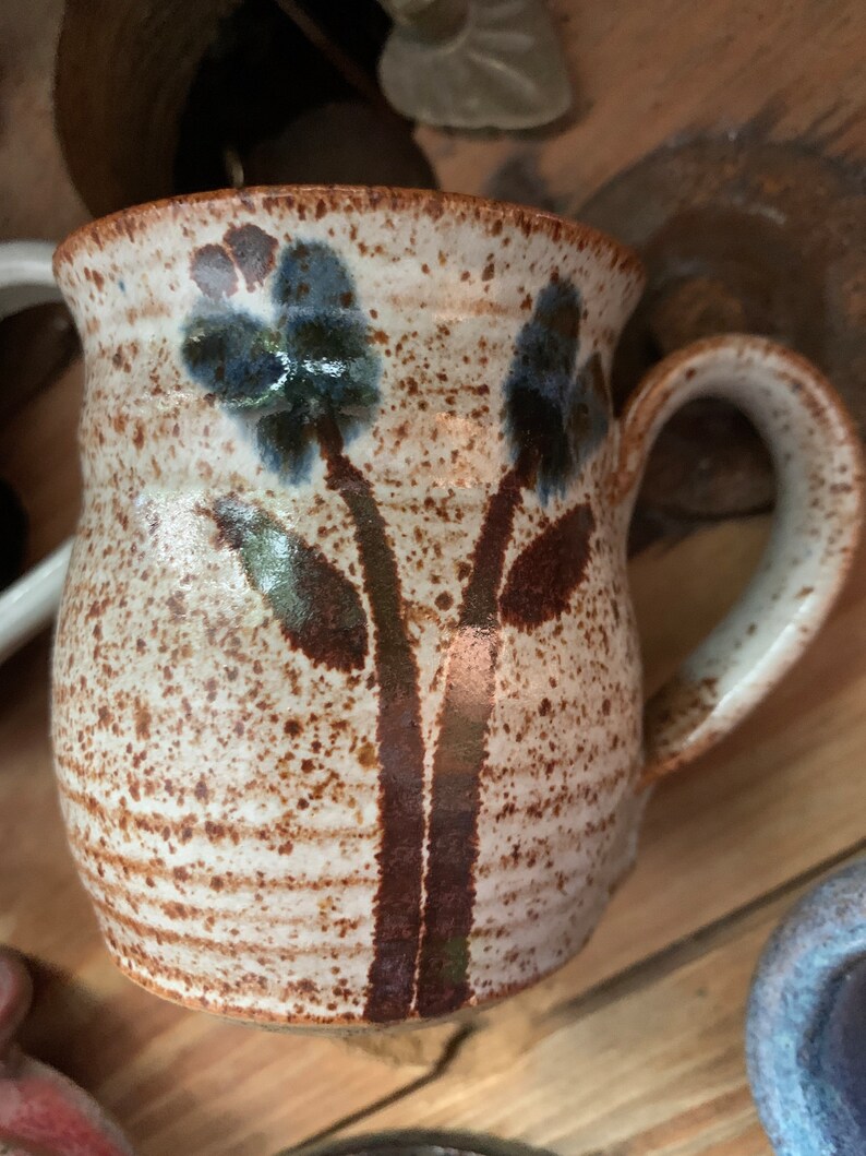 以下が含まれることがあります： 茶色の斑点のある釉薬と青い花がついた茶色の花の模様が施された白い陶器のマグカップ。マグカップは丸みを帯びたボディとハンドルが付いています。