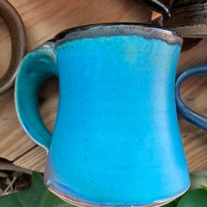 May include: A turquoise ceramic mug with a black rim and a slightly flared shape. The mug has a handle that curves out from the side.