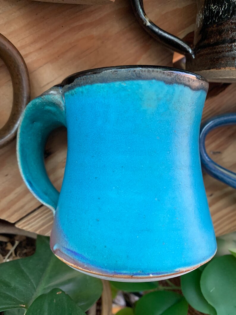 May include: A turquoise ceramic mug with a black rim and a slightly flared shape. The mug has a handle that curves out from the side.