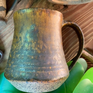 May include: A rustic, brown and green ceramic mug with a speckled glaze. The mug has a wide base and a rounded handle.