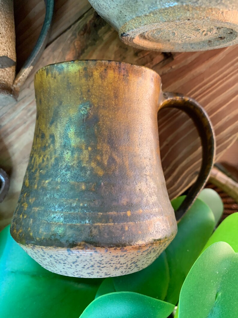May include: A rustic, brown and green ceramic mug with a speckled glaze. The mug has a wide base and a rounded handle.