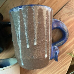May include: A brown ceramic mug with a blue handle and white glaze dripping down the sides. The mug has a speckled texture.