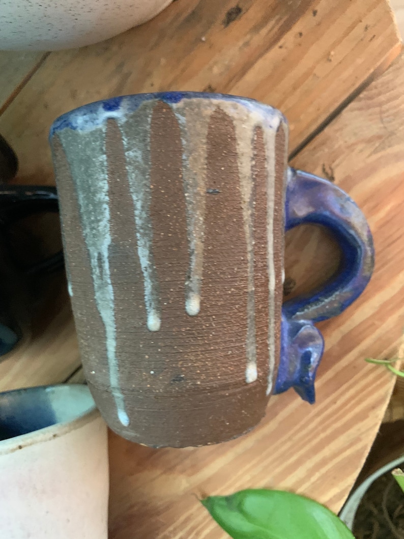 May include: A brown ceramic mug with a blue handle and white glaze dripping down the sides. The mug has a speckled texture.