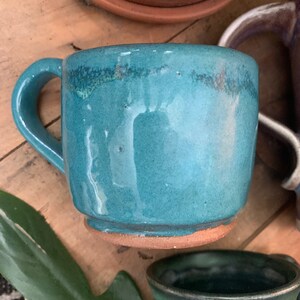 May include: A teal blue ceramic mug with a brown rim and base. The mug has a rounded shape and a handle.