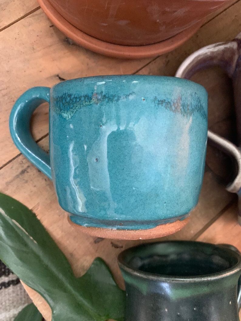 May include: A teal blue ceramic mug with a brown rim and base. The mug has a rounded shape and a handle.