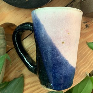 May include: A ceramic mug with a black handle and a blue and pink glaze. The mug has a unique, slightly uneven shape.