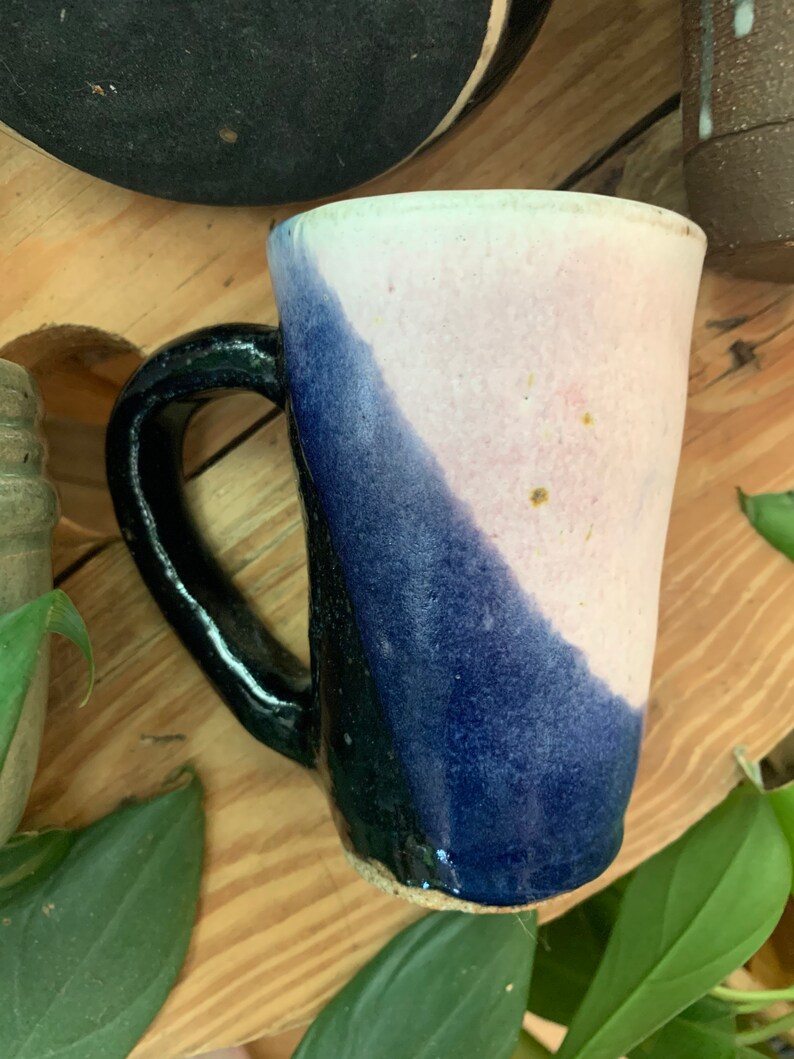 May include: A ceramic mug with a black handle and a blue and pink glaze. The mug has a unique, slightly uneven shape.