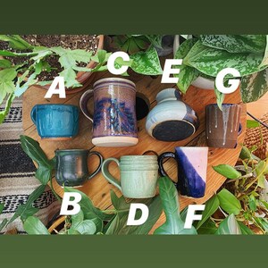 May include: A collection of seven ceramic mugs and vases in various colours and patterns, including blue, green, brown, and purple. The mugs and vases are arranged on a wooden surface with green plants in the background.