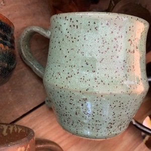 May include: A speckled green ceramic mug with a brown handle. The mug has a rounded body and a speckled glaze.