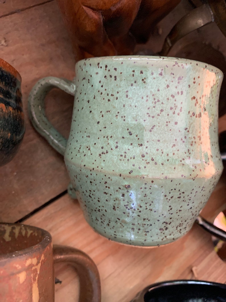 May include: A speckled green ceramic mug with a brown handle. The mug has a rounded body and a speckled glaze.