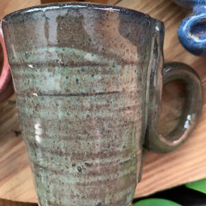 以下が含まれることがあります： 茶色と緑の釉薬をかけた手作りセラミックマグカップ。マグカップは、ユニークでわずかに非対称なハンドルを持っています。
