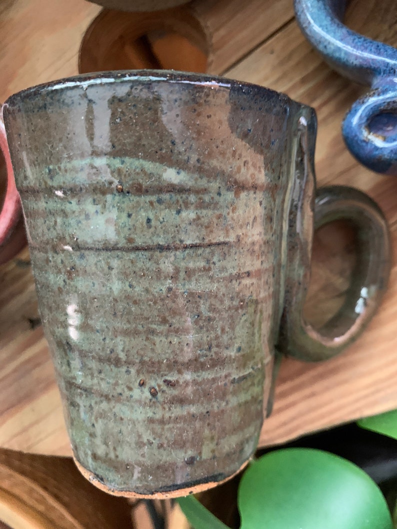 以下が含まれることがあります： 茶色と緑の釉薬をかけた手作りセラミックマグカップ。マグカップは、ユニークでわずかに非対称なハンドルを持っています。