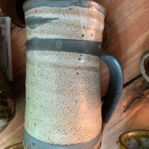 May include: A handmade ceramic mug with a white glaze and gray stripes. The mug has a handle and a rounded bottom.