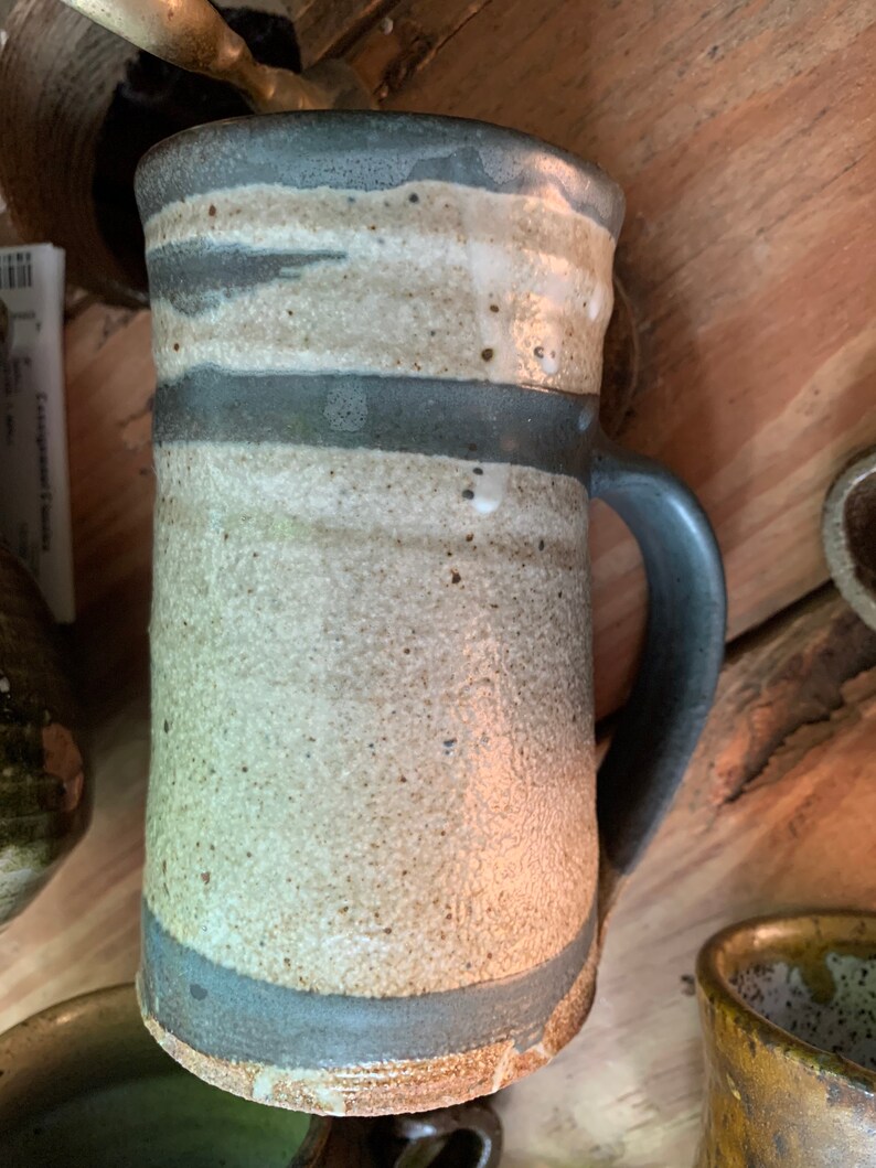 May include: A handmade ceramic mug with a white glaze and gray stripes. The mug has a handle and a rounded bottom.