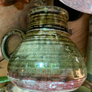 May include: A green and brown ceramic pitcher with a rounded body and a handle. The pitcher has a dark brown glaze with streaks of green and a red rim.