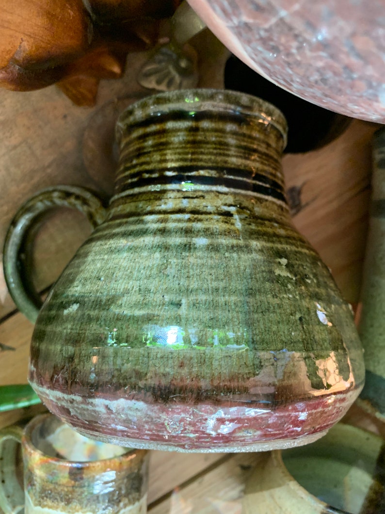 May include: A green and brown ceramic pitcher with a rounded body and a handle. The pitcher has a dark brown glaze with streaks of green and a red rim.