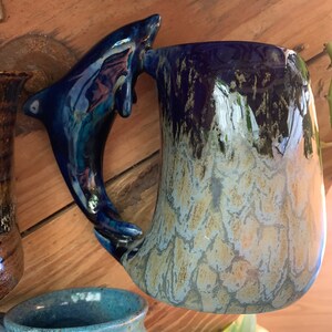 May include: A blue and white ceramic mug with a dolphin-shaped handle. The mug has a speckled glaze and a light blue and white colour gradient.