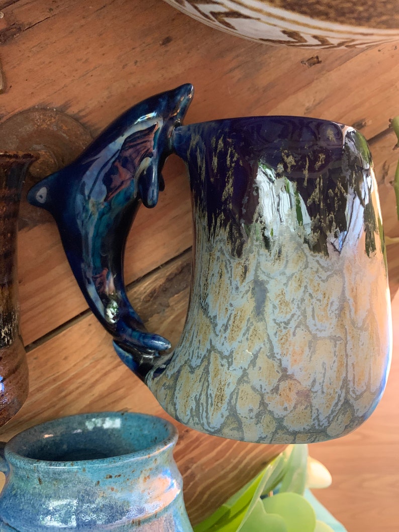 May include: A blue and white ceramic mug with a dolphin-shaped handle. The mug has a speckled glaze and a light blue and white colour gradient.