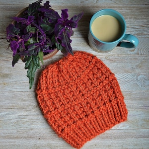Könnte beinhalten: Eine leuchtend orangefarbene Strickmütze auf einer Holzoberfläche. Die Mütze ist aus einem dicken Garn gefertigt und hat ein schlichtes, klassisches Design.