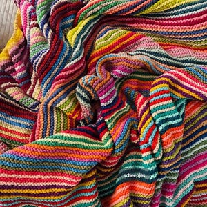May include: A hand-knitted blanket featuring a vibrant, multi-colored striped pattern with horizontal rows in shades of red, orange, yellow, green, blue, and purple. The fabric has a distinct garter stitch texture, and the word scheepjes is visible.