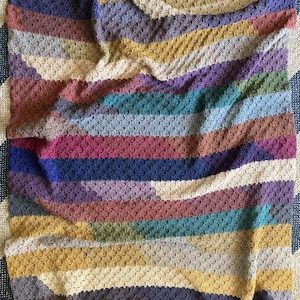 May include: A multi-coloured crocheted blanket with a striped pattern. The blanket is made with a variety of colours, including mustard yellow, teal, purple, and brown.